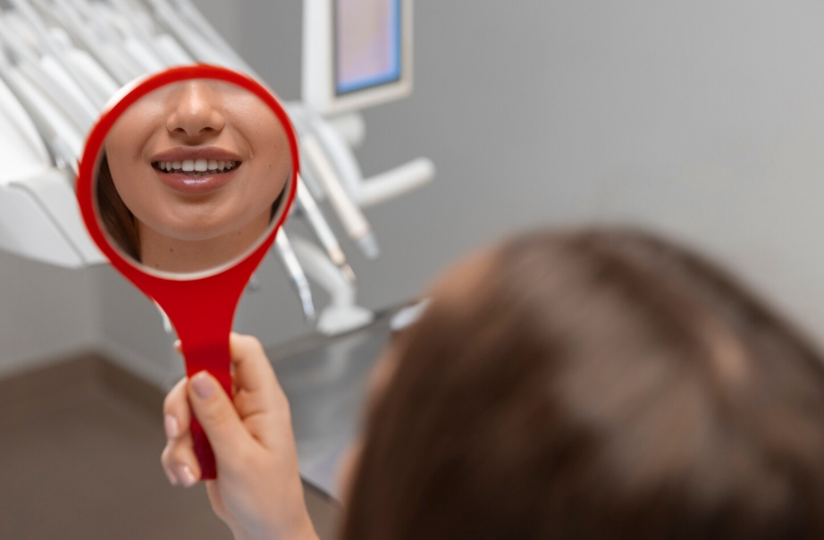 Young woman smiling while holding a dental mirror reflecting her beautiful teeth during a clinic visit.