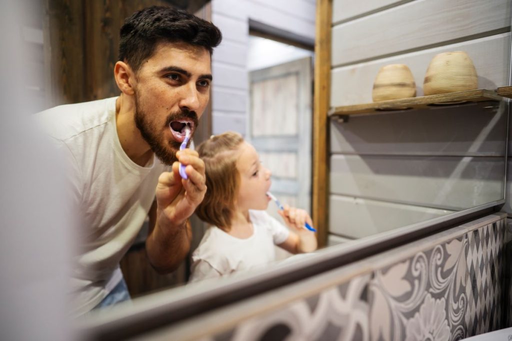 Father and daughter brushing their teeth together to maintain oral health for all ages and lifelong care at home.
